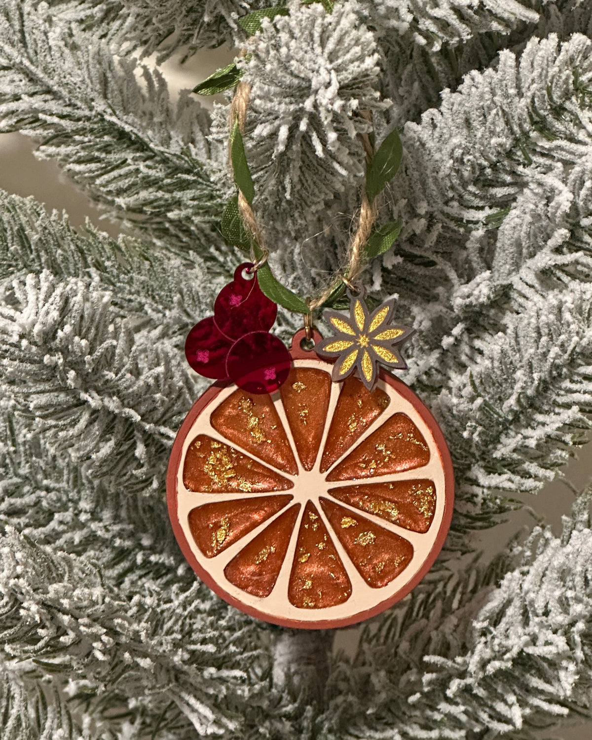 A wooden dried orange slice Christmas ornament with gold glitter details, hanging on a flocked  Christmas tree. Ornament comes with acrylic cranberry and cardamom charms.