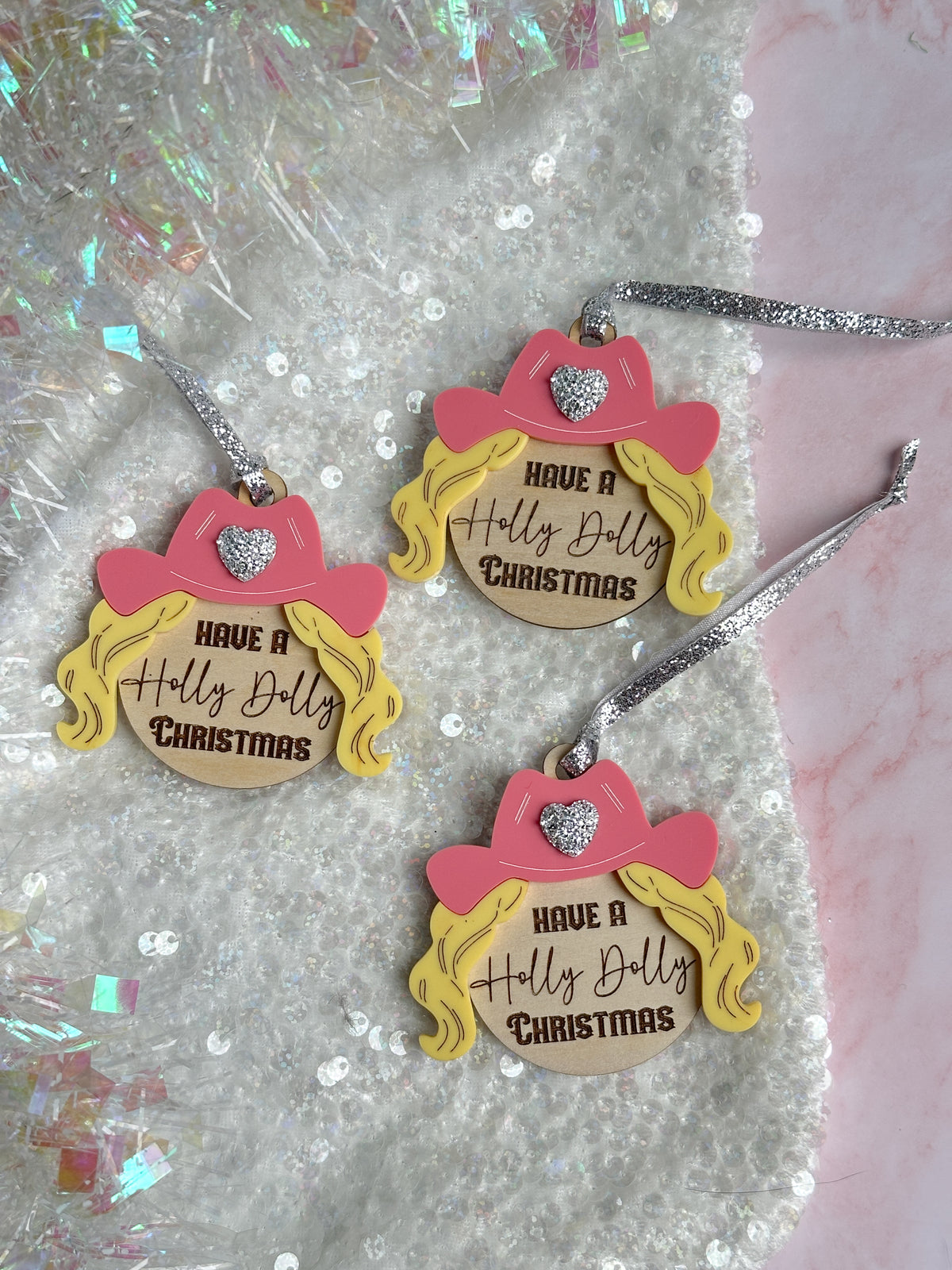 Three matching Christmas ornaments shaped like cartoon girl profiles with pink hats, yellow hair, and silver hanging ribbons, each printed with the phrase 'HAVE A Holly Dolly Christmas,' arranged on a sequined white surface with pink and iridescent tinsel accents.
