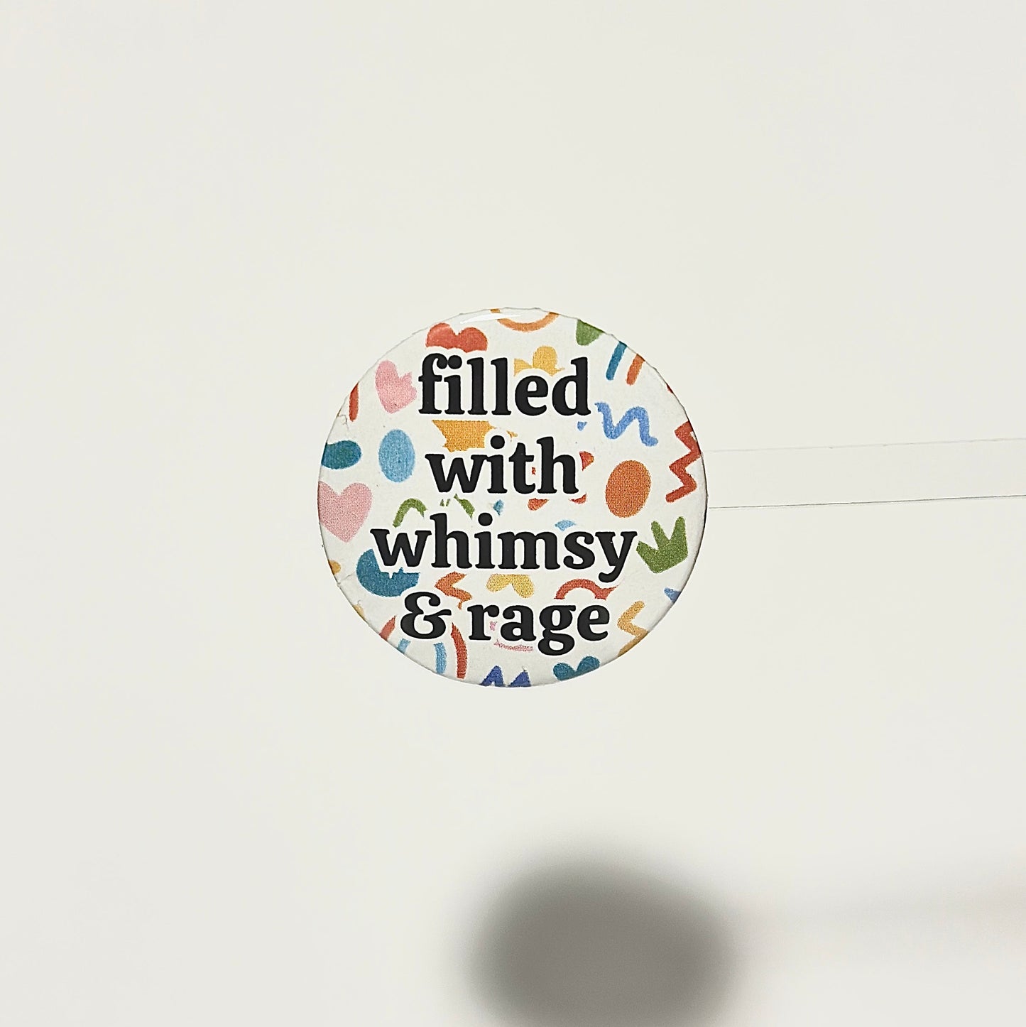 Round sticker with text 'filled with whimsy & rage' on a white background with abstract multicolor shapes