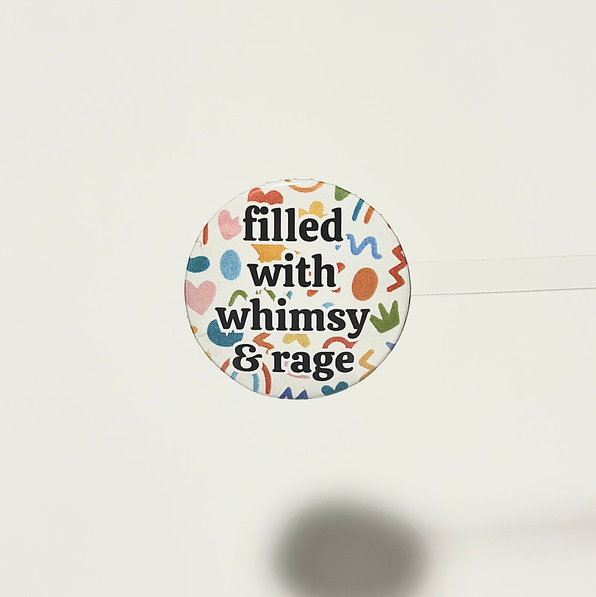 Round sticker with text 'filled with whimsy & rage' on a white background with abstract multicolor shapes